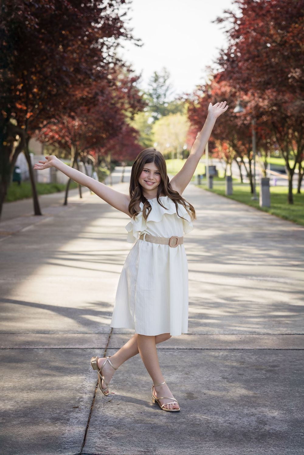 Person in a white dress jumping playfully on a tree-lined path with autumn foliage.