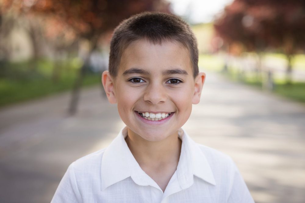 A series of seven headshots showing a young boy in a white collared shirt smiling against a blurred outdoor background.