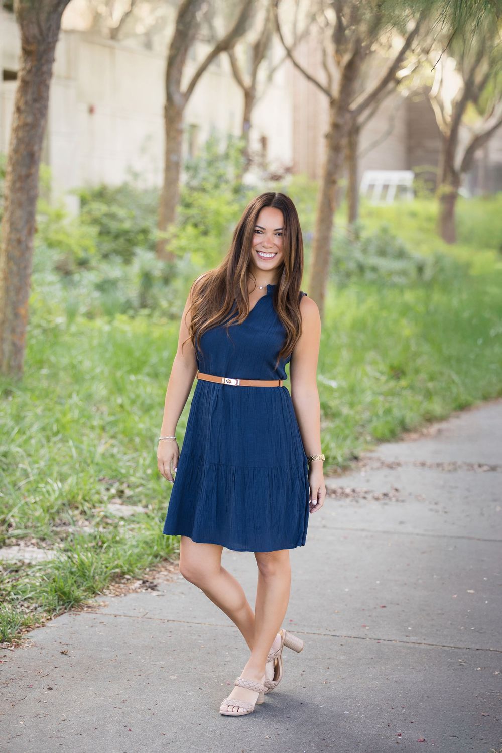 Person in a navy blue dress poses playfully on a tree-lined path.