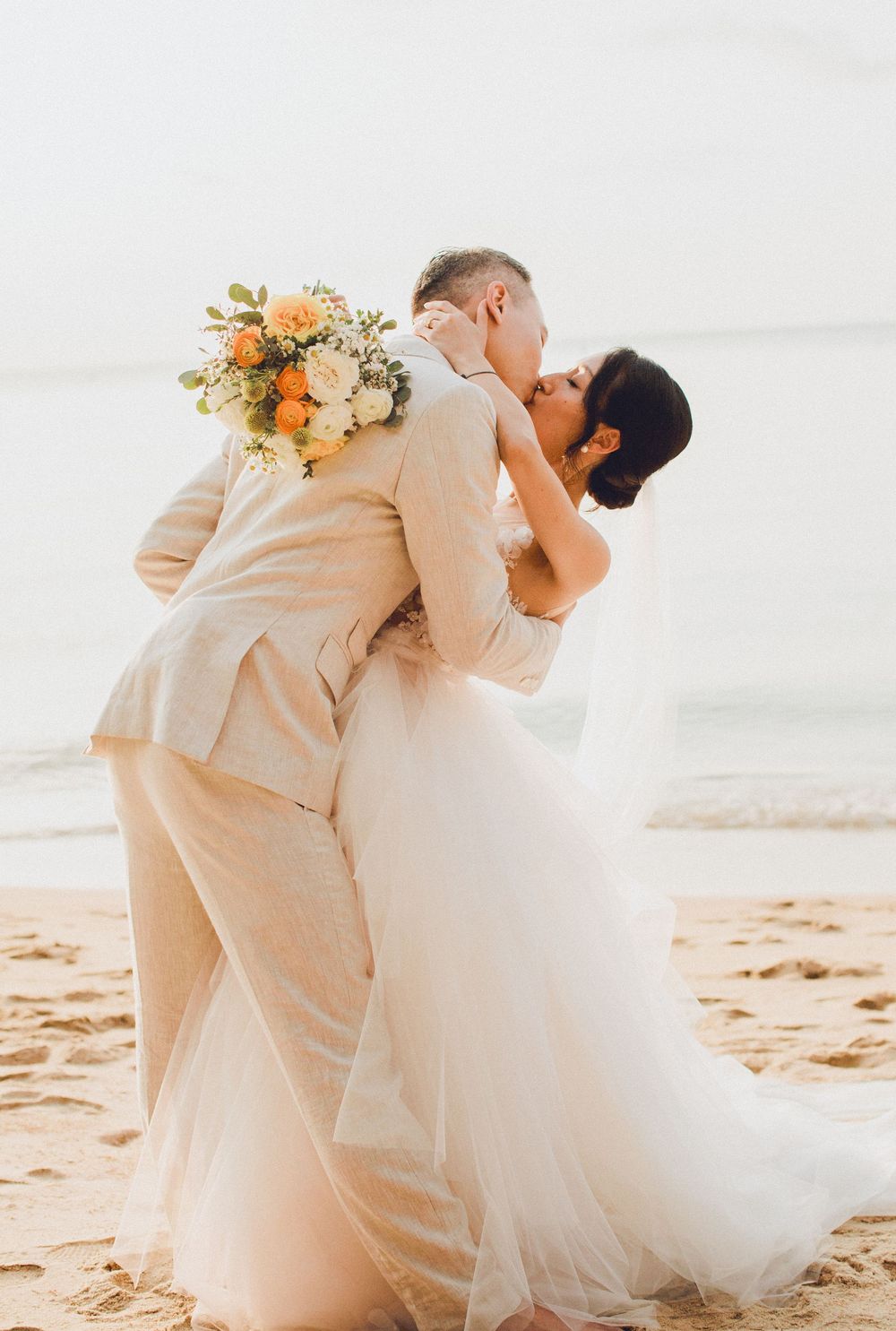 Groom dips and kisses bride on a Koh Samui beach, holding bouquet behind his back in a romantic wedding moment.