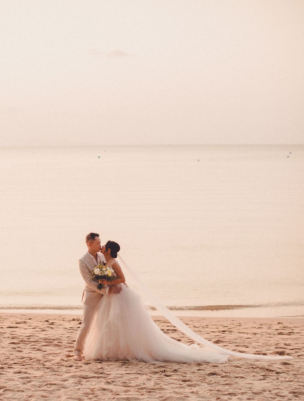 Bride and groom embracing on a sandy Koh Samui beach with flowing veil during sunset wedding photos