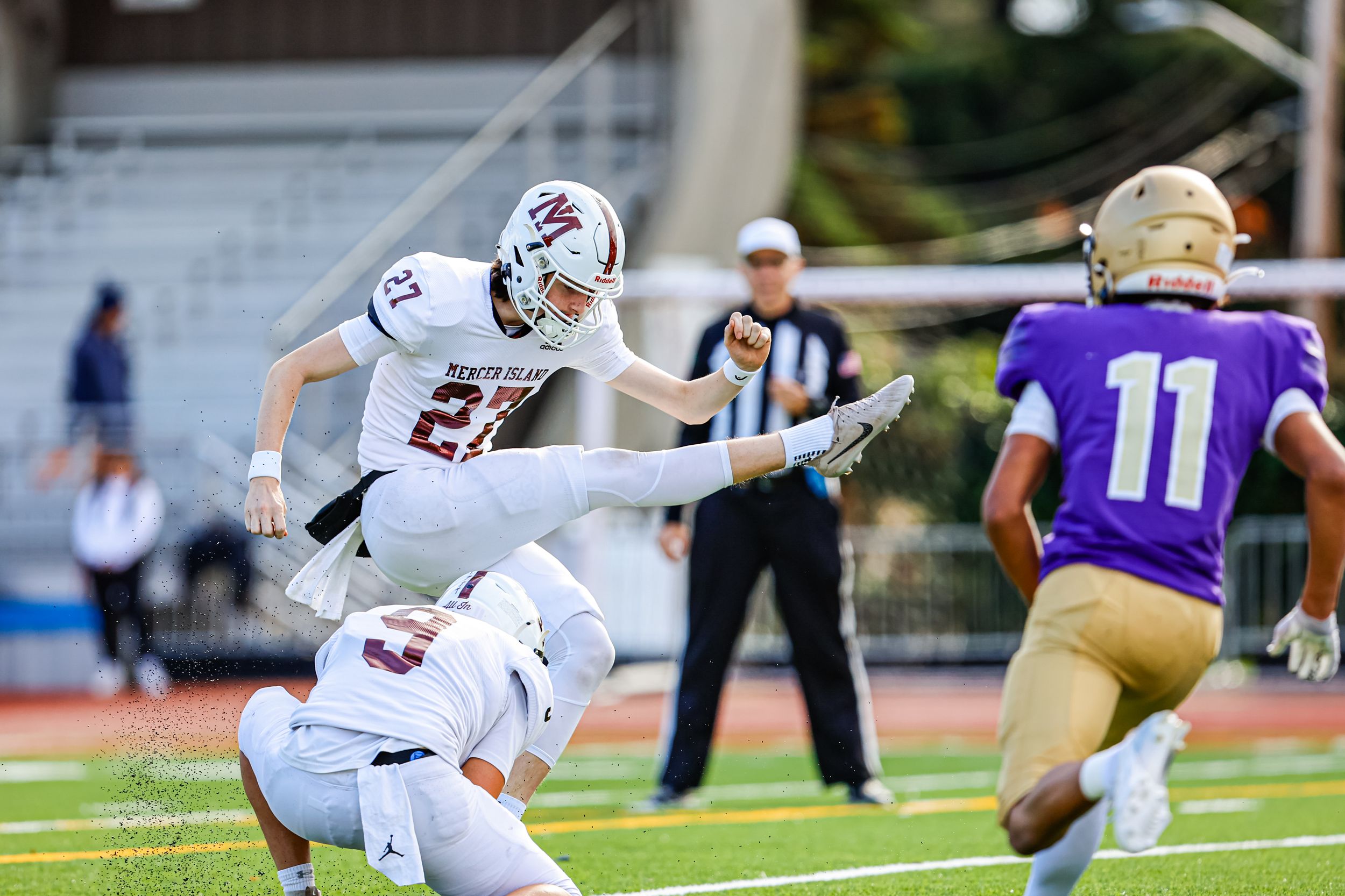 Football player #21 leaps over a fallen teammate during a high school football game on a sunny day.