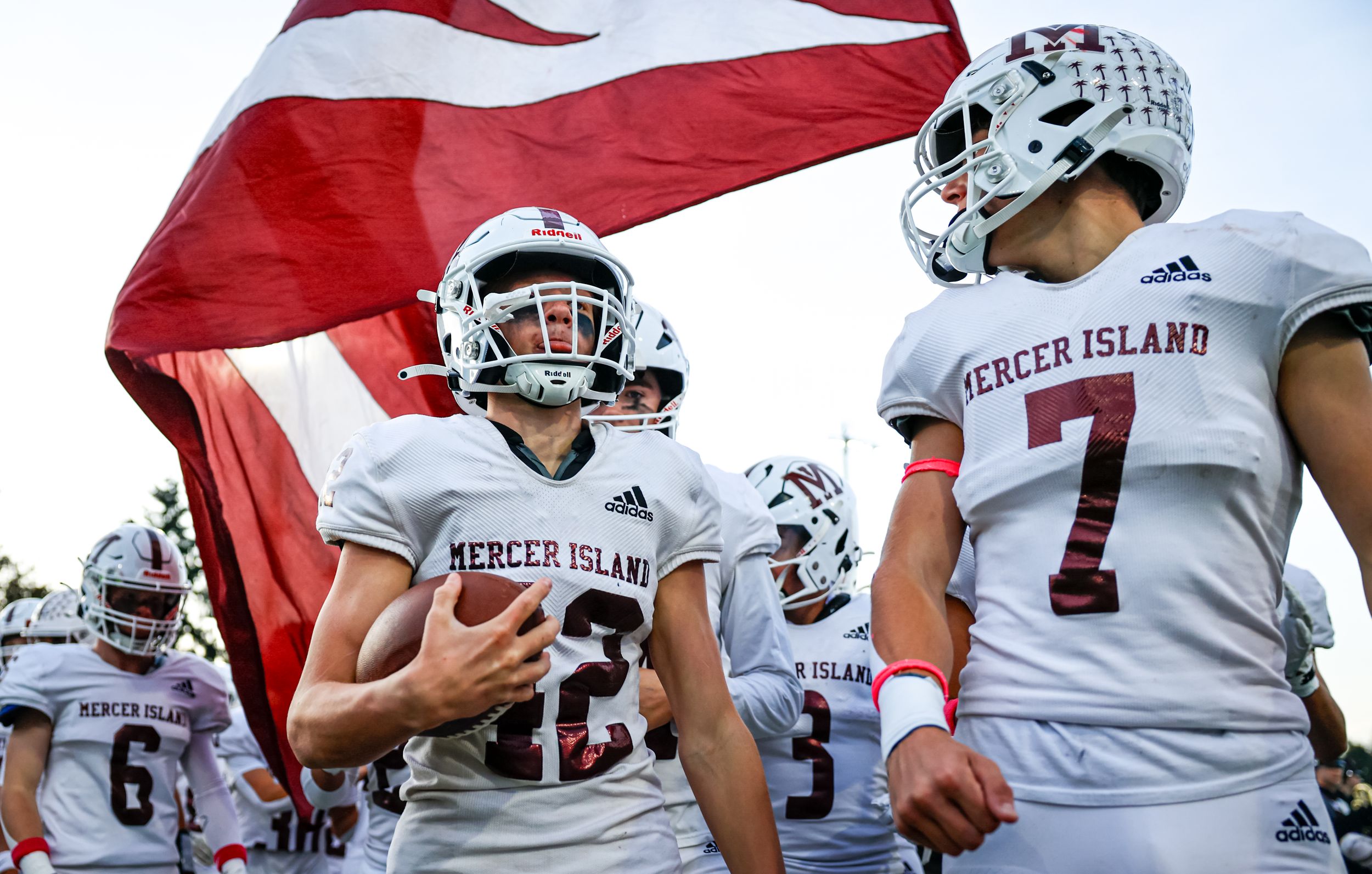 Mercer Island football players in white uniforms take the field under a red and white flag during a game.