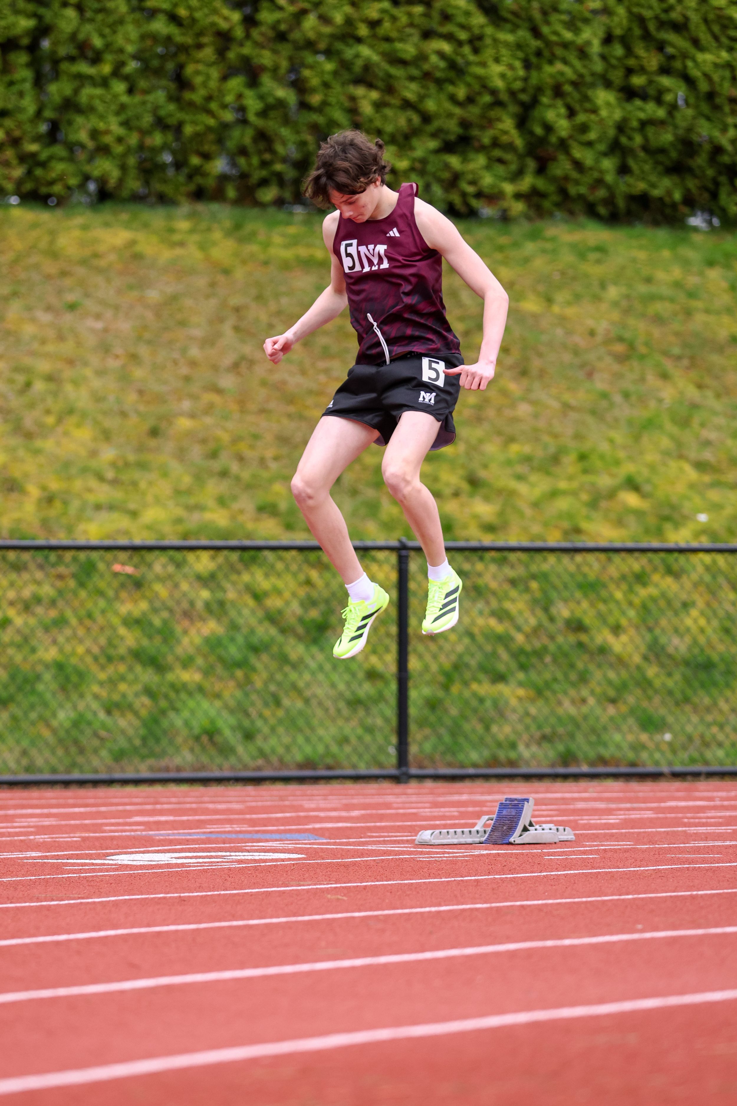 Young male track athlete in maroon uniform leaping over hurdle on red track during outdoor competition.