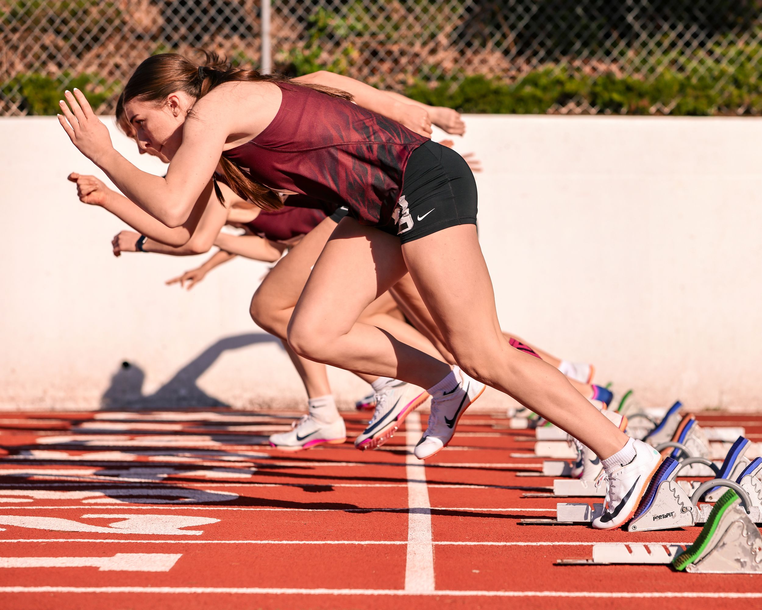 Female track athletes explode from starting blocks on a red running track during a sprint race competition.