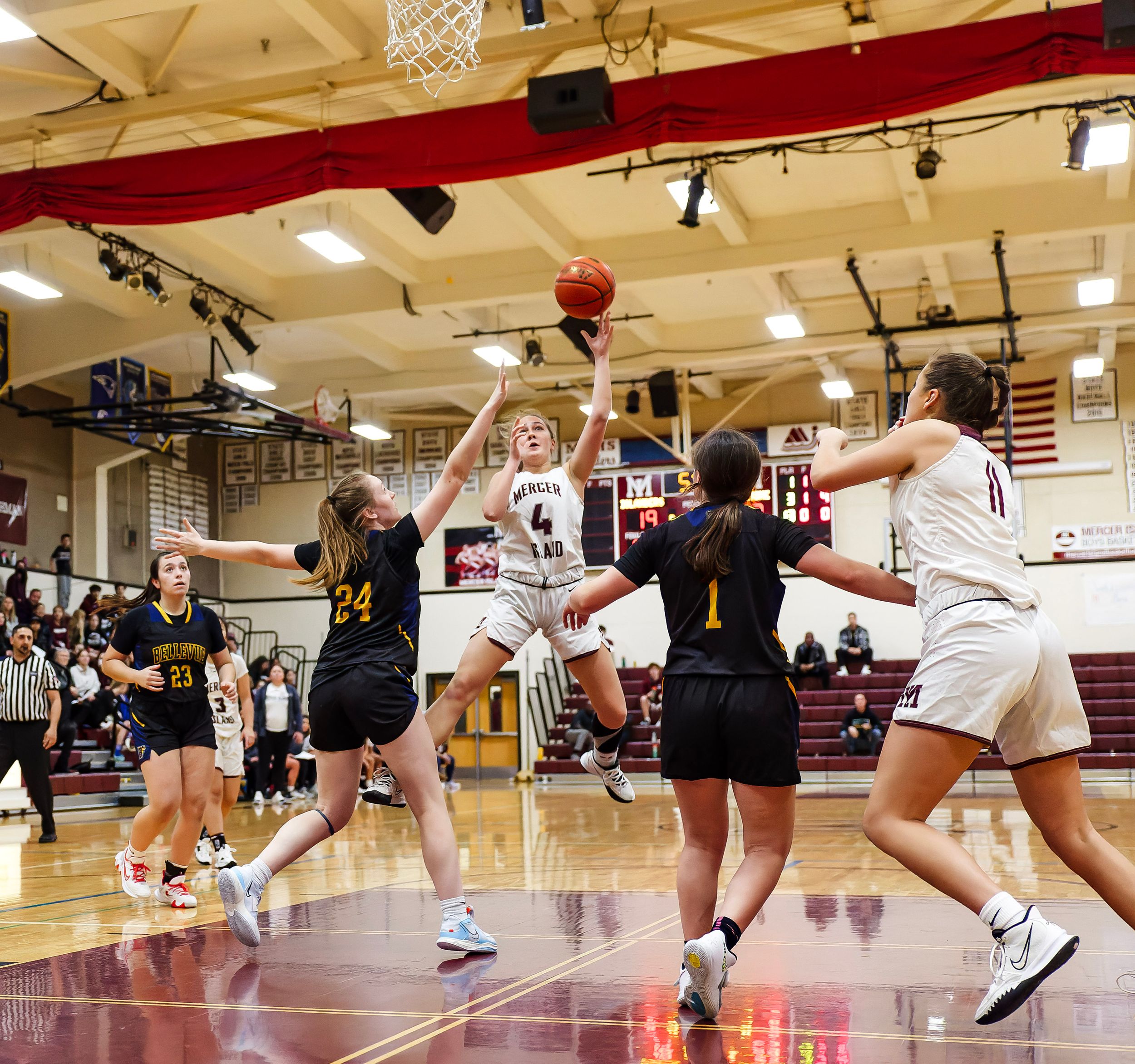 Girls high school basketball player leaps for a layup shot during an intense indoor game with players in maroon and black uniforms.