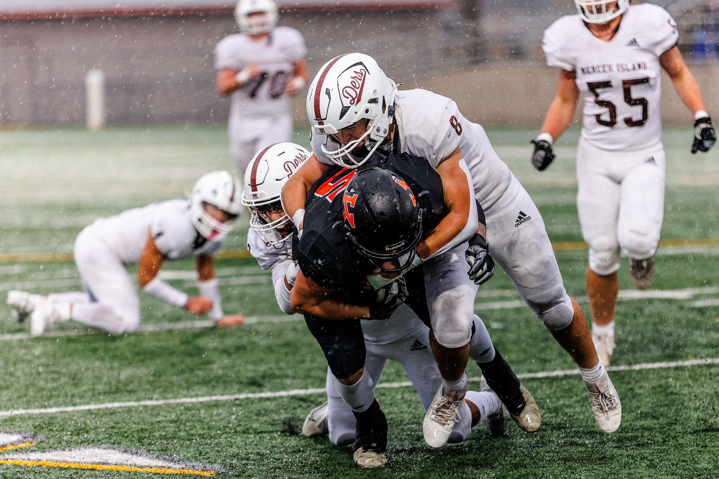 Football players tackle opponent during a rainy high school game, with players wearing white and dark uniforms on field.