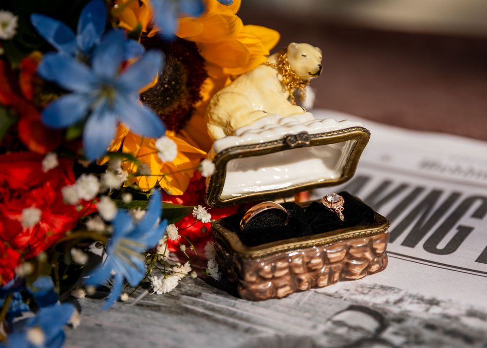 Vintage jewelry box with two gold rings beside colorful flowers on a newspaper backdrop.