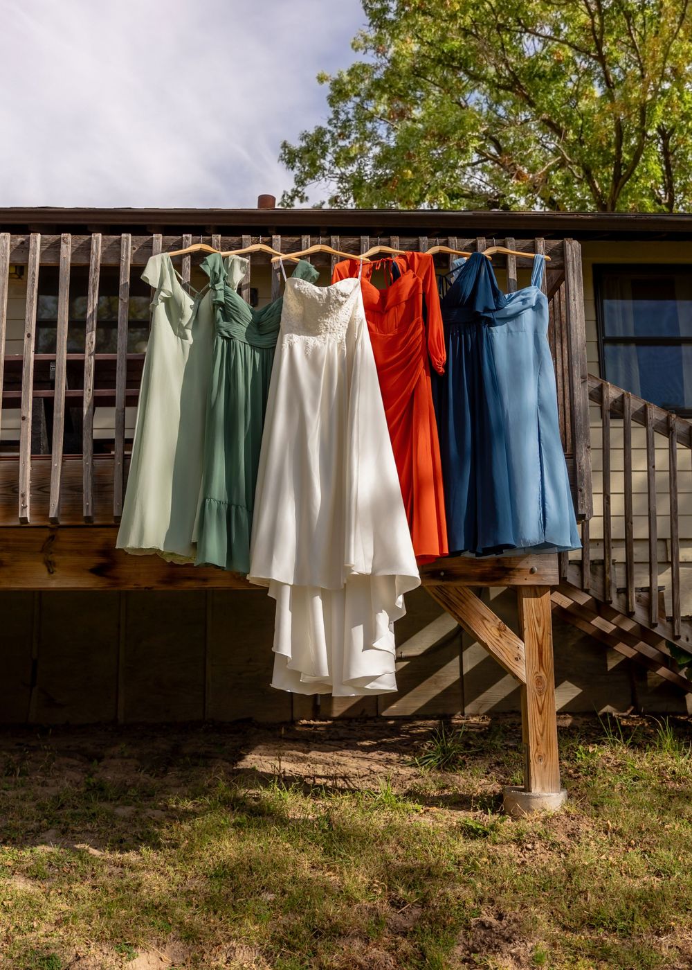 Colorful bridesmaid dresses and white wedding gown hanging outdoors on a wooden deck railing  at the Cottonwood at Platte River State Park