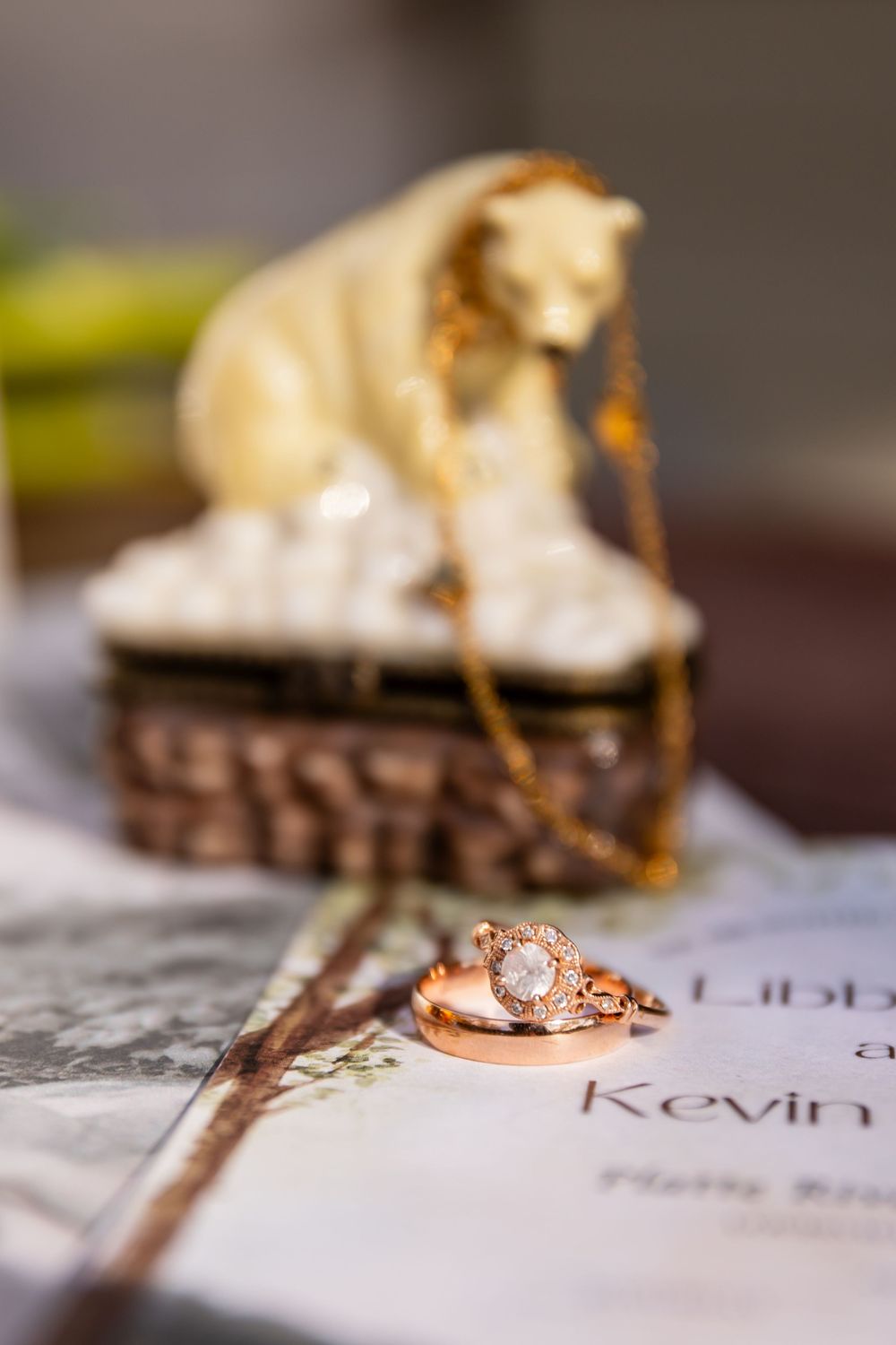 Close-up of rose gold wedding rings and necklace on wedding invitation next to a decorative bear figurine jewelry box.