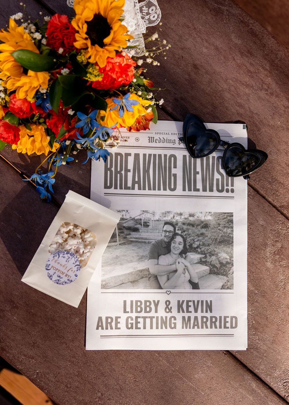 Wedding announcement newspaper for Libby & Kevin with sunflowers, sunglasses, and a couple photo on a wooden table.