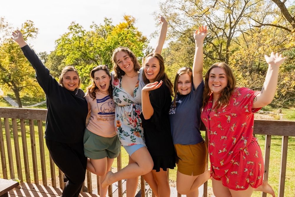 Six bridesmaids celebrating on a wooden deck with arms raised surrounded by autumn trees.