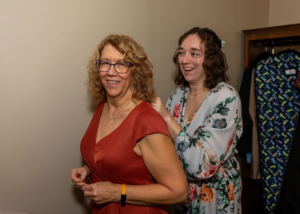 Two women smiling and laughing together at an indoor social event, one in red dress, other in floral top.