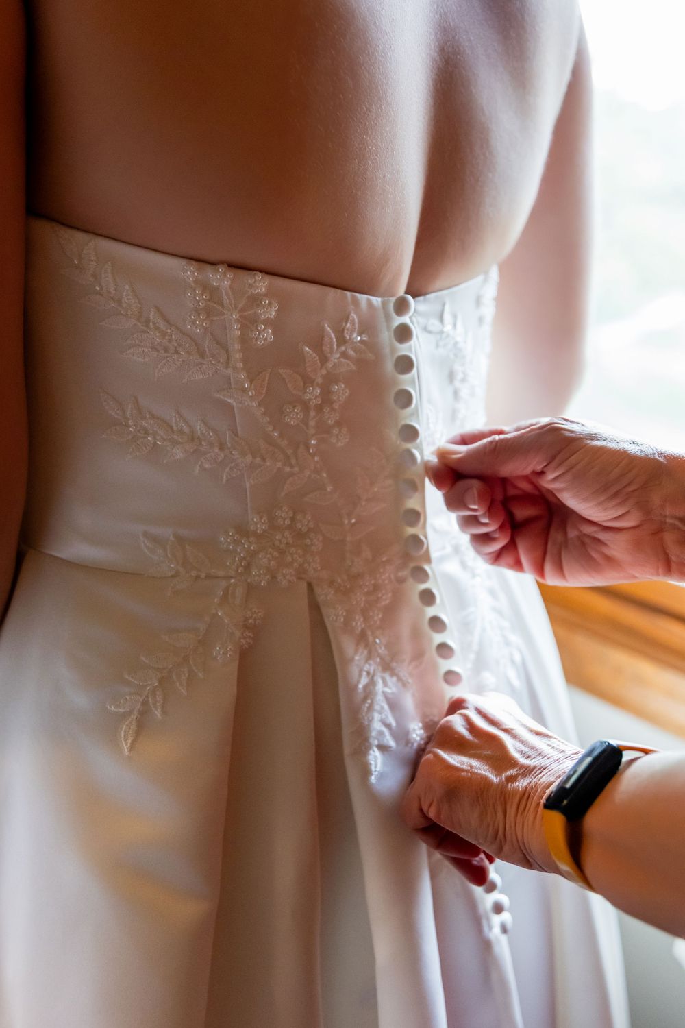 Hands buttoning the back of a white lace wedding dress with floral embroidery details.