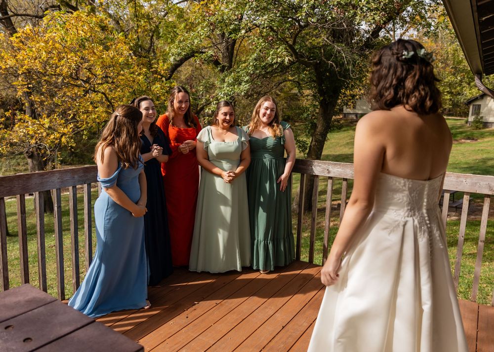 Bridesmaids in blue, red, and green gowns reacting to bride's first look reveal on outdoor deck.