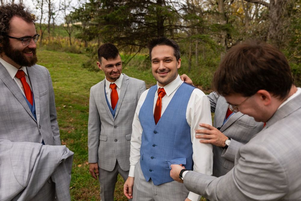 Groom smiling in blue vest and red tie with groomsmen in light gray suits outdoors.