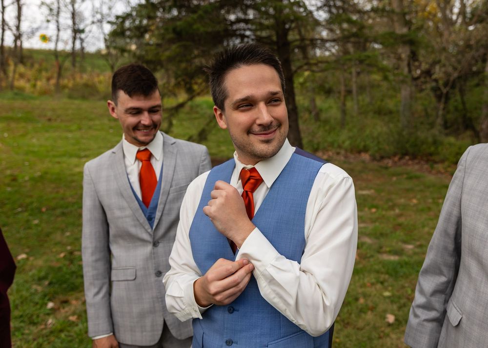 Groom in blue vest and red tie adjusting his cufflinks outdoors with groomsmen nearby.