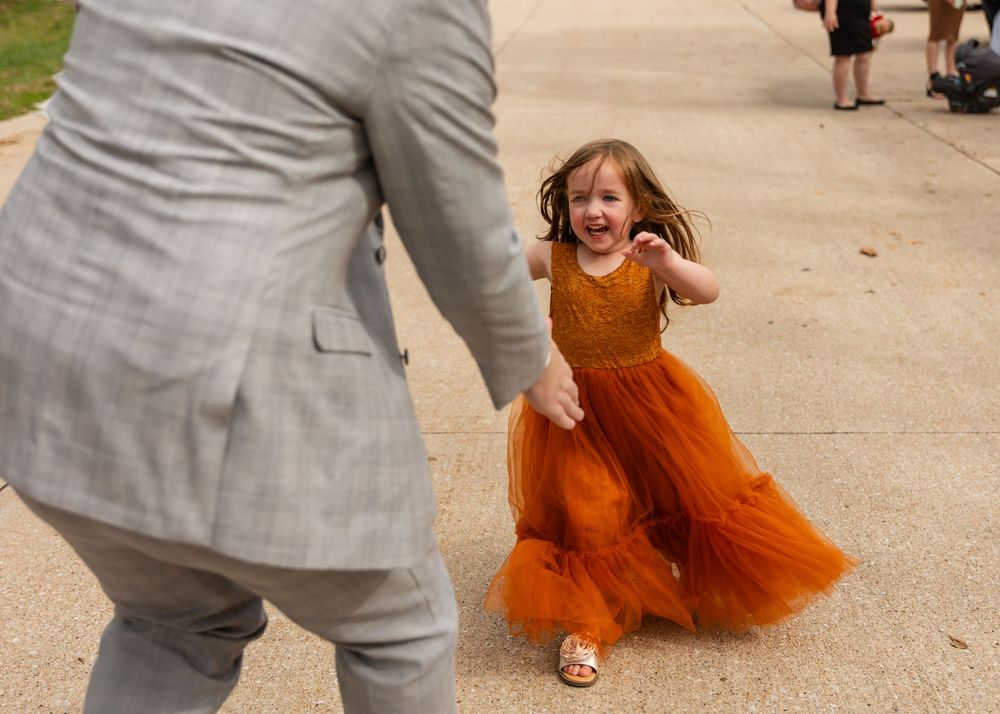 Excited little girl in orange tulle dress running toward person in gray suit at wedding.