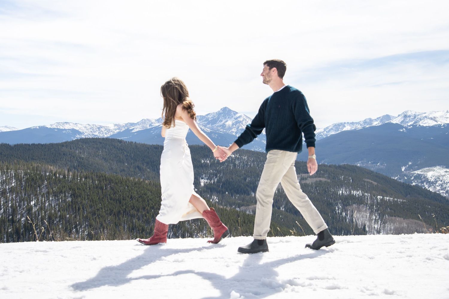 Chic fashionable couple holding hands walking in snow with mountain landscape backdrop for Vail Colorado winter engagement photos.