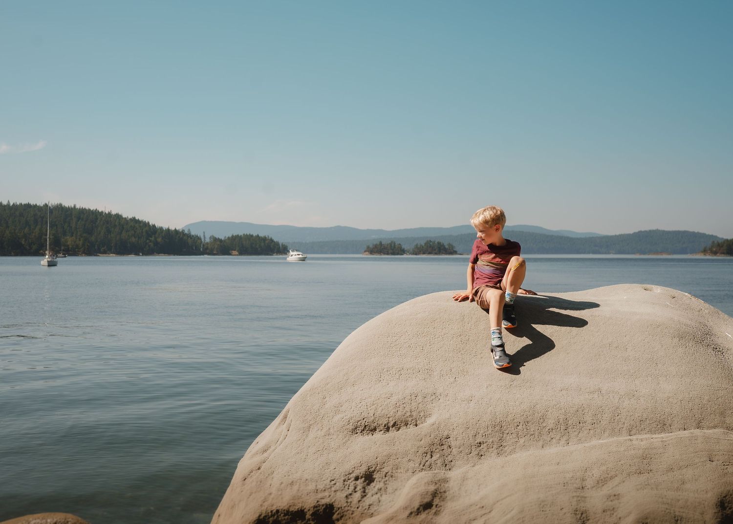 Child sitting on a large rock by a calm lake with forested islands and mountains in the background on a sunny day.