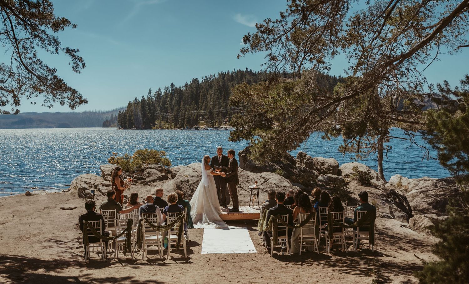 Intimate outdoor wedding ceremony on rocky lakeshore with seated guests beneath pine trees.