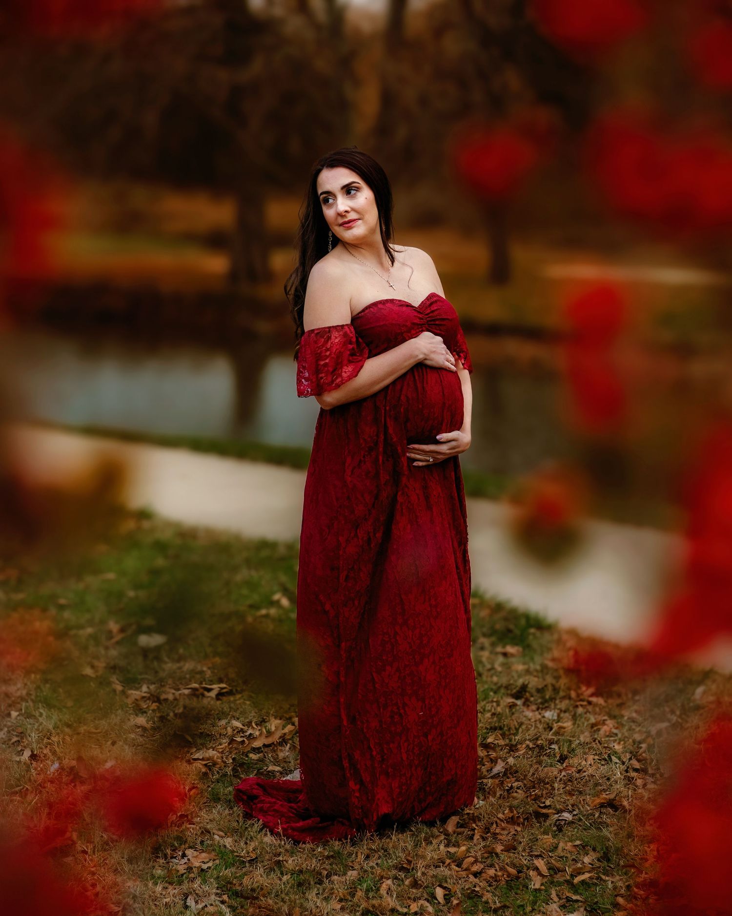 A pregnant woman in a flowing red lace gown poses outdoors with autumn foliage and bokeh effects in the background in Colleyville, TX.