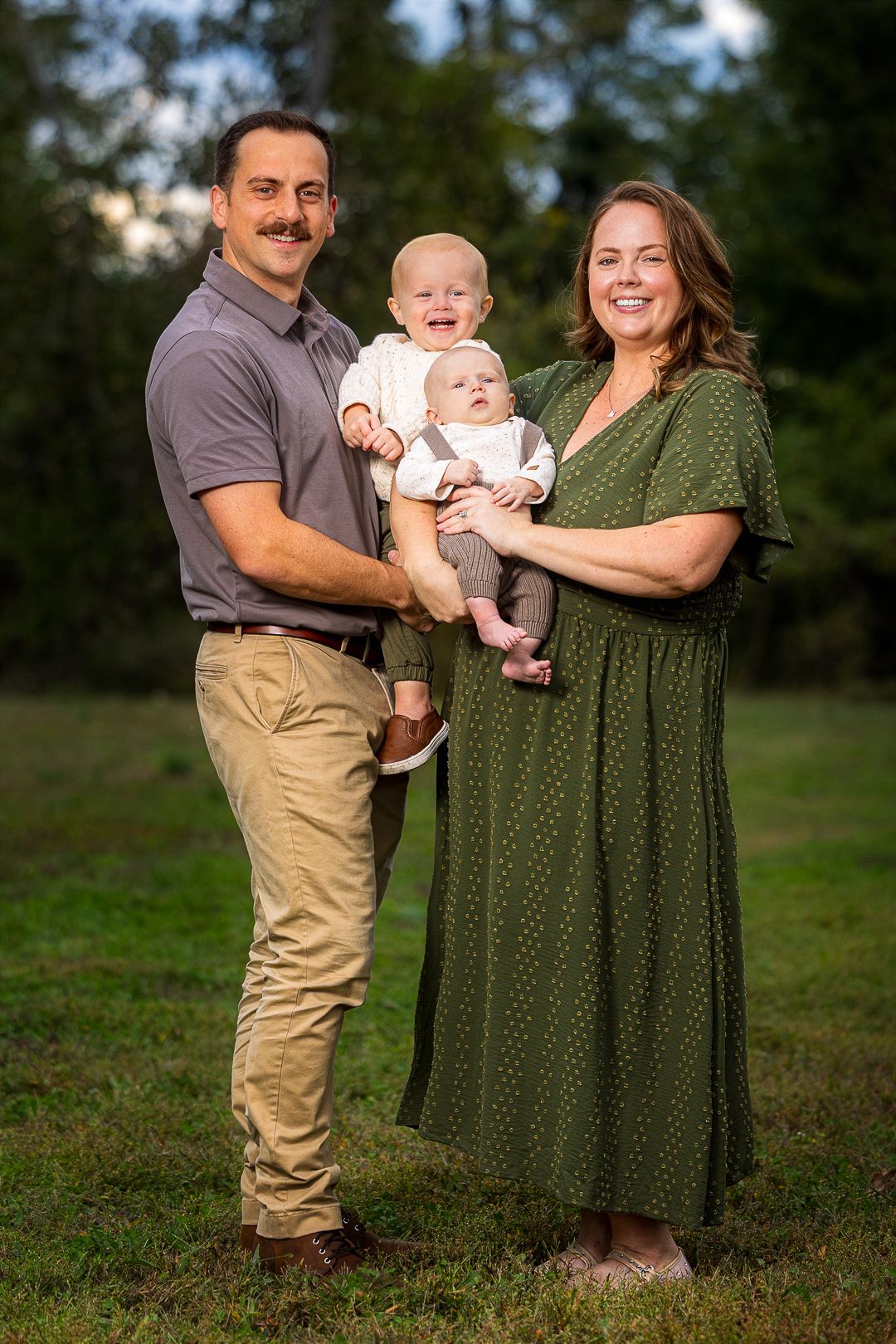 A family in casual attire poses together outdoors on grass with a small baby wearing white clothing.