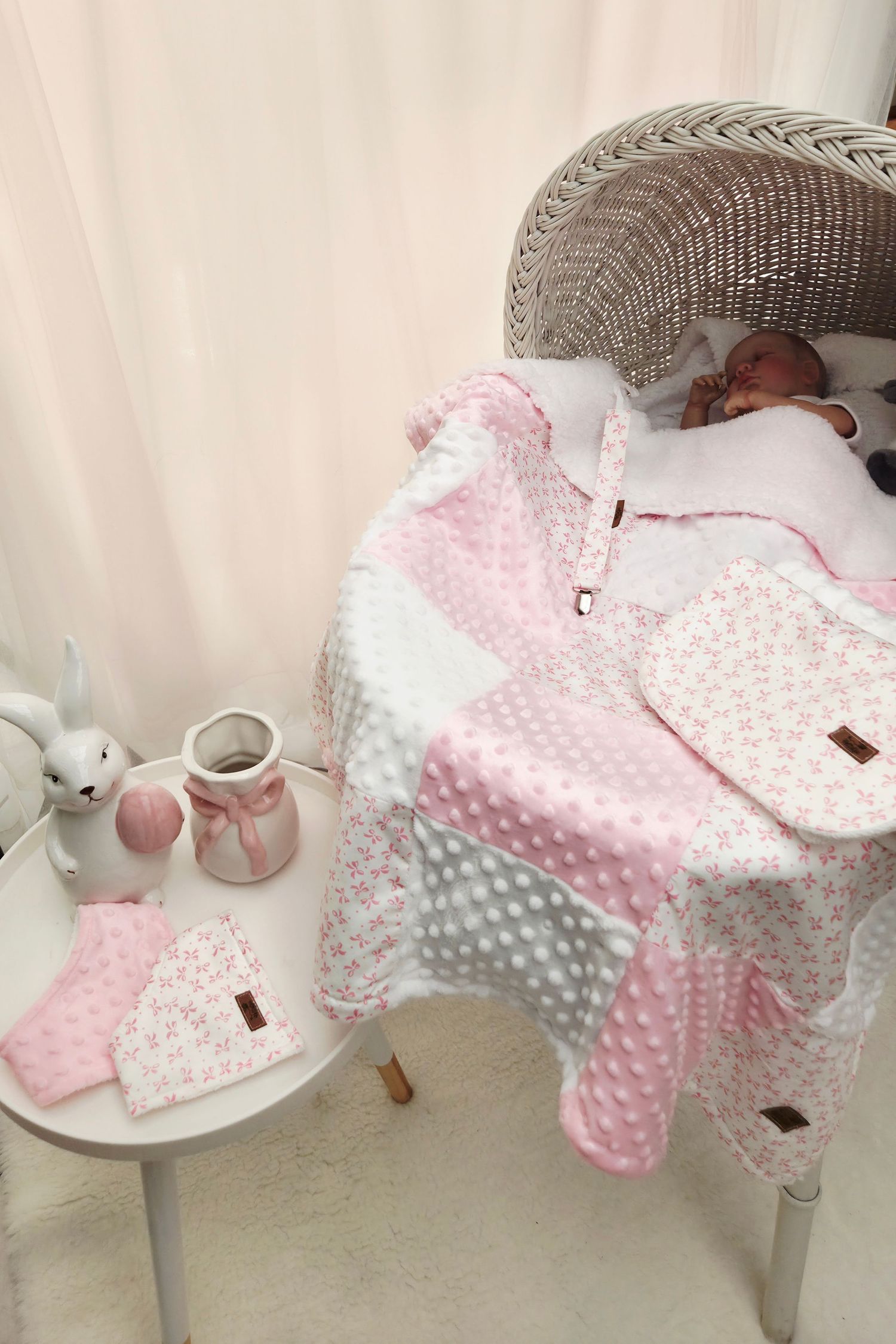 Newborn baby resting on a pink patchwork quilt blanket in a wicker chair with matching booties and accessories nearby.