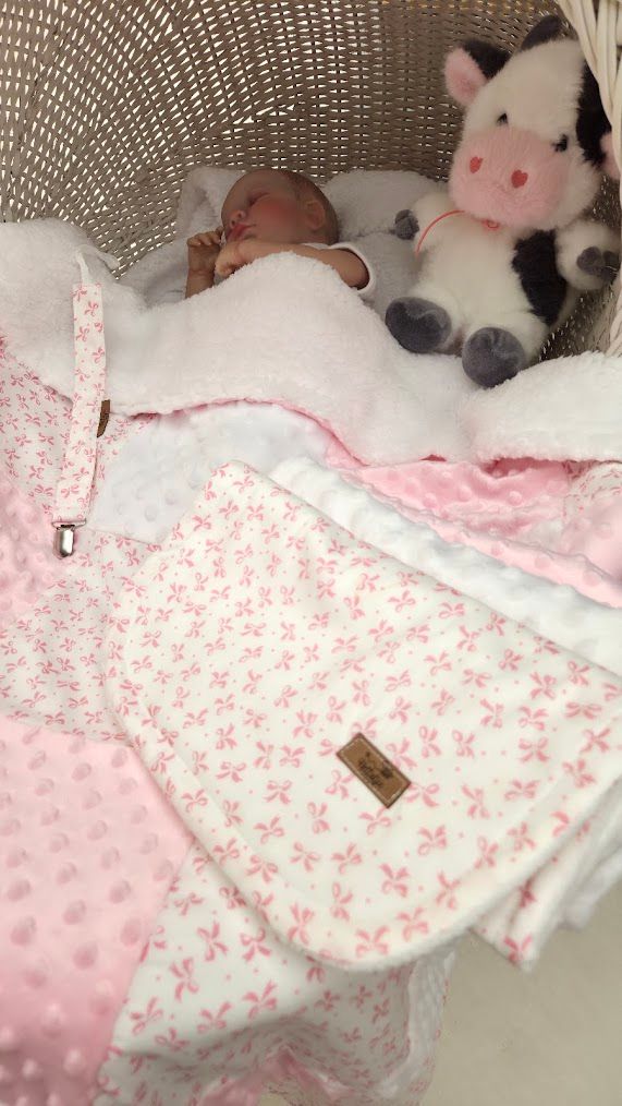 Newborn baby sleeping in a wicker bassinet with a pink floral blanket and a black and white cow stuffed animal toy.