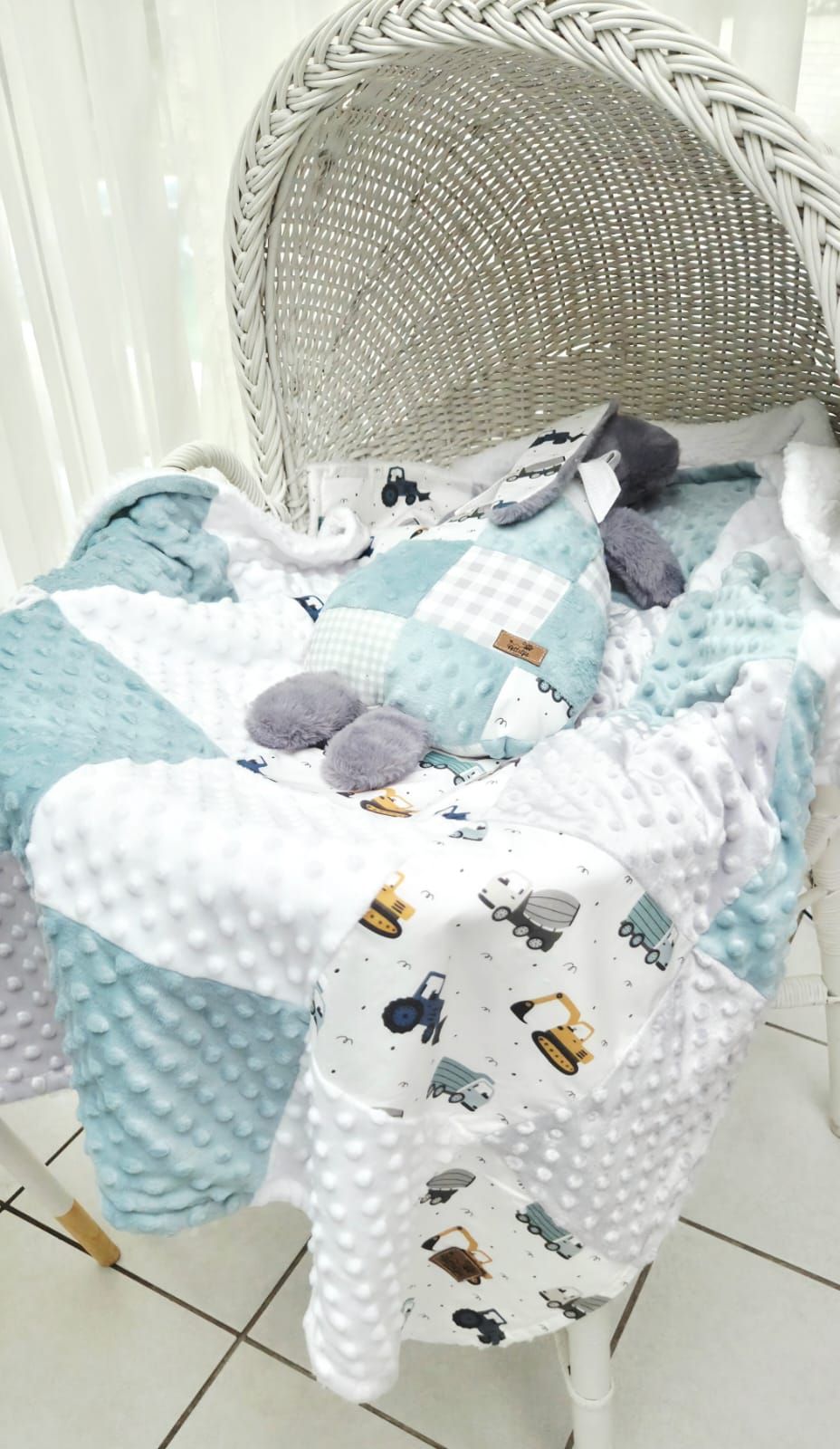 Baby quilt set in mint, white, and gray with construction vehicle print, displayed on a white wicker chair.