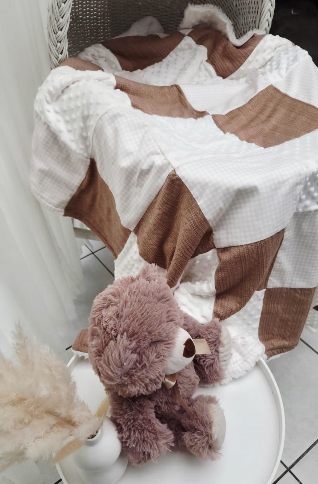 Brown and white checkered baby blanket on a wicker chair with a fluffy mauve teddy bear on a white side table.