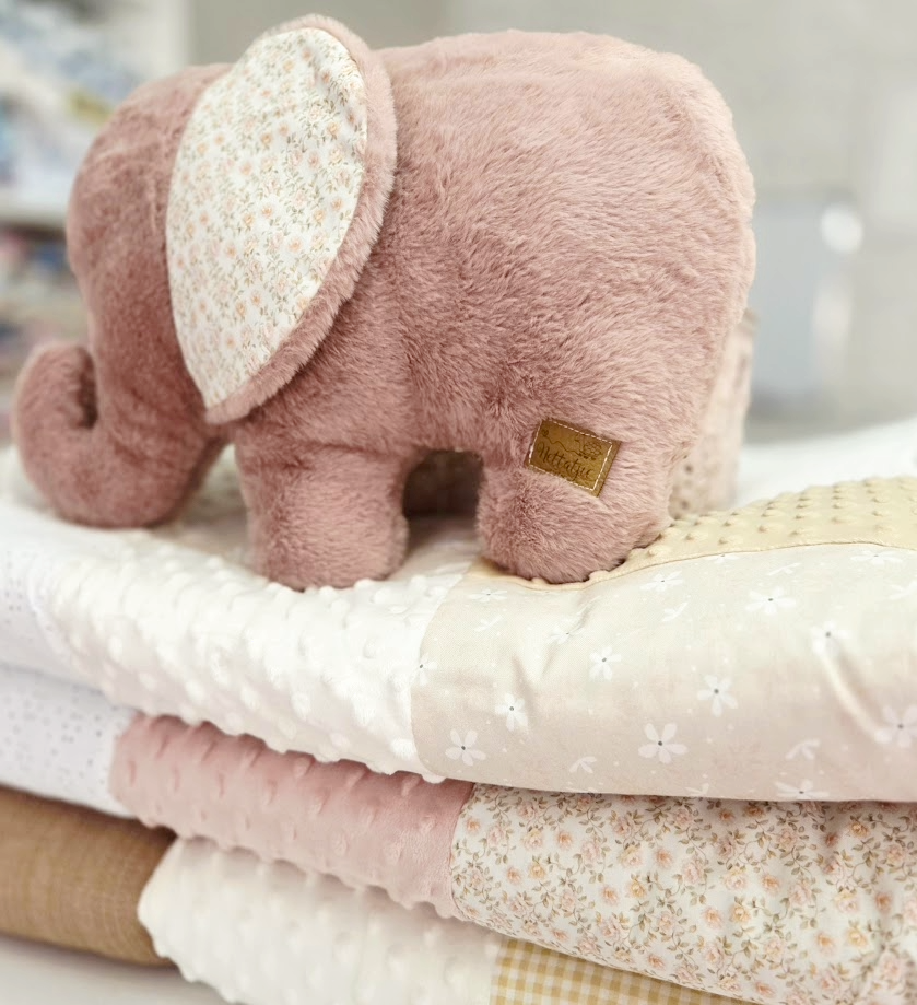 A soft pink plush elephant toy sitting on top of neatly folded pastel baby blankets in floral and dotted patterns.