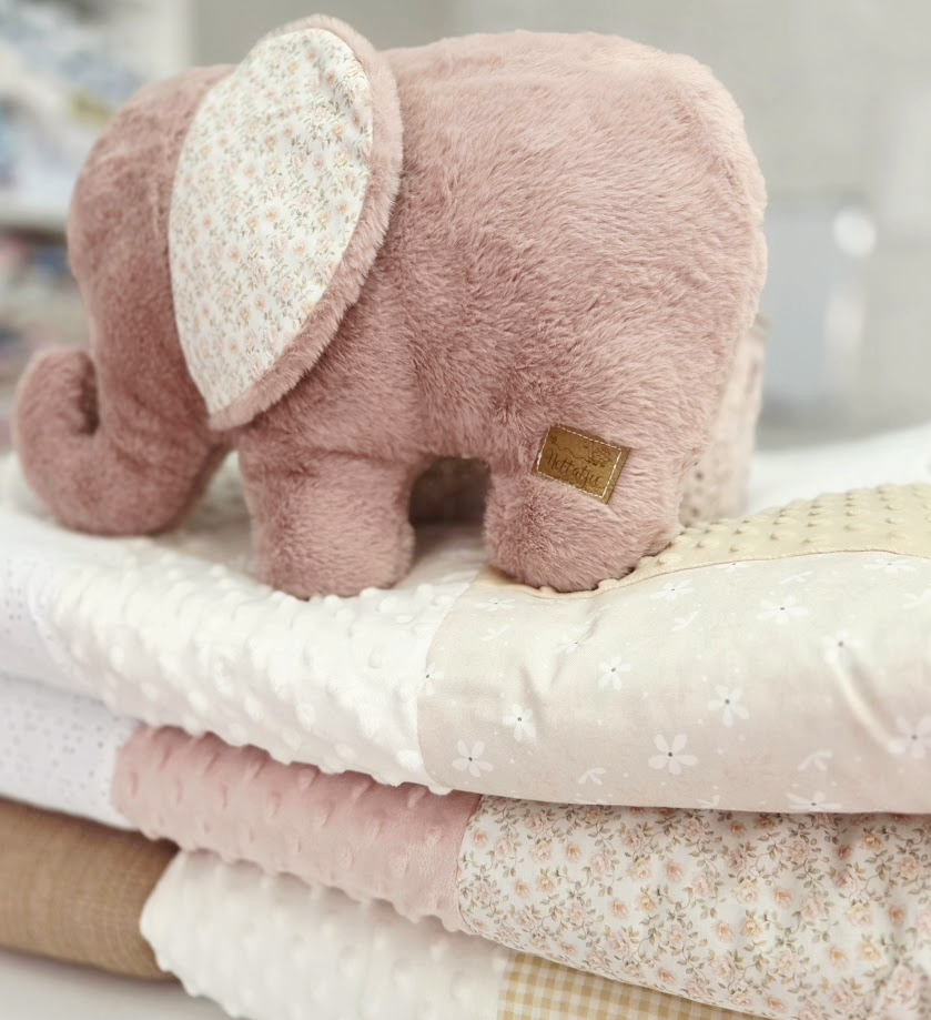 A plush pink elephant toy resting on a stack of soft folded baby blankets in pastel pink and beige tones.