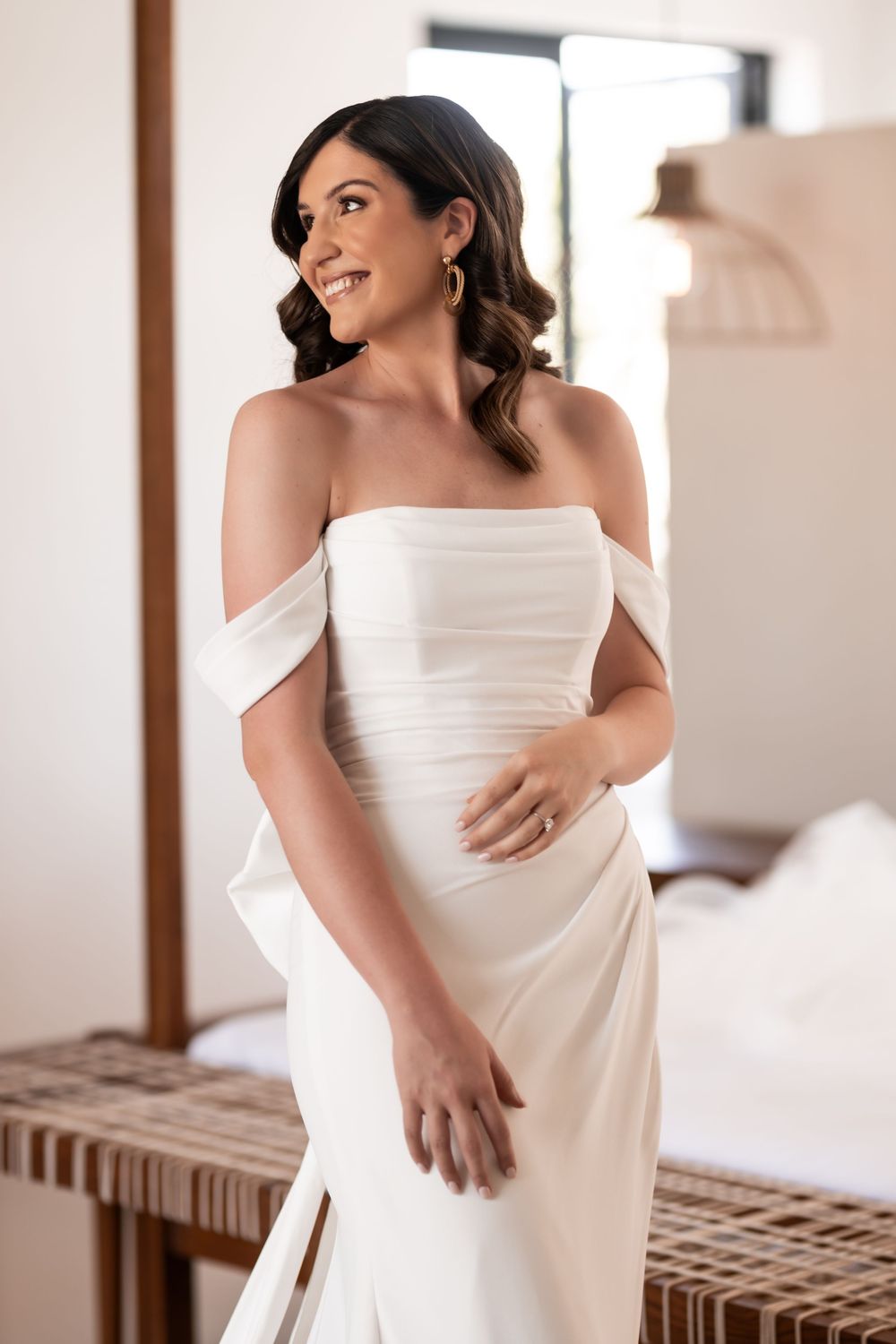 Smiling bride in elegant off-shoulder white satin wedding gown posing indoors with soft natural lighting.
