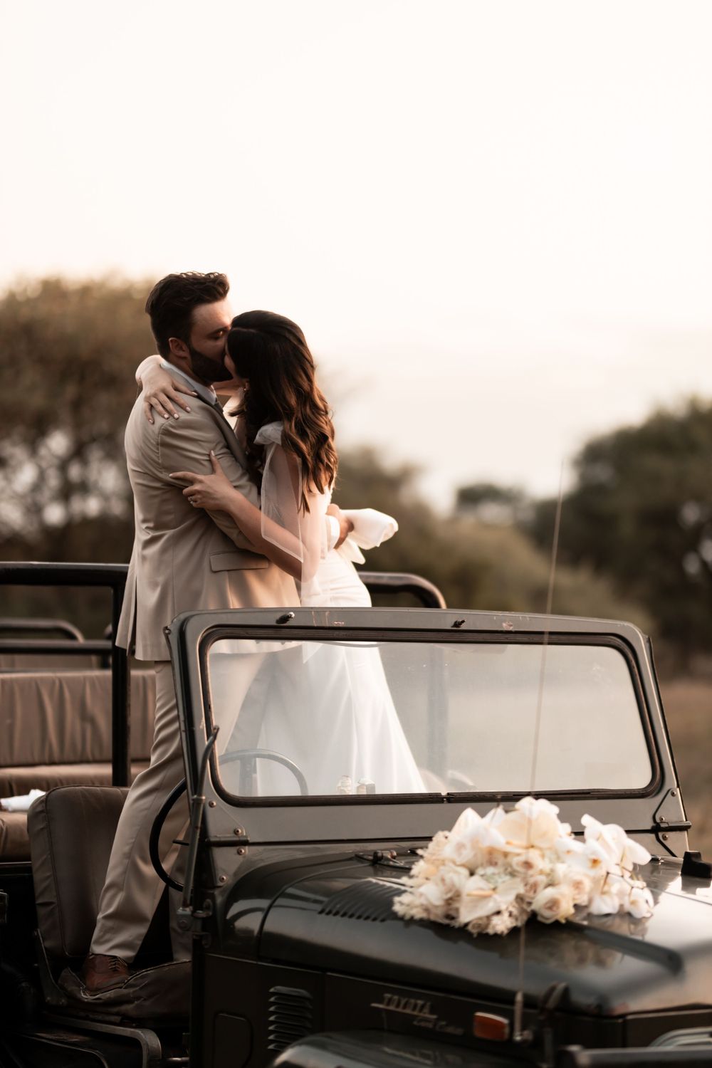 Bride and groom share a romantic kiss beside a vintage safari jeep adorned with white flowers at sunset.