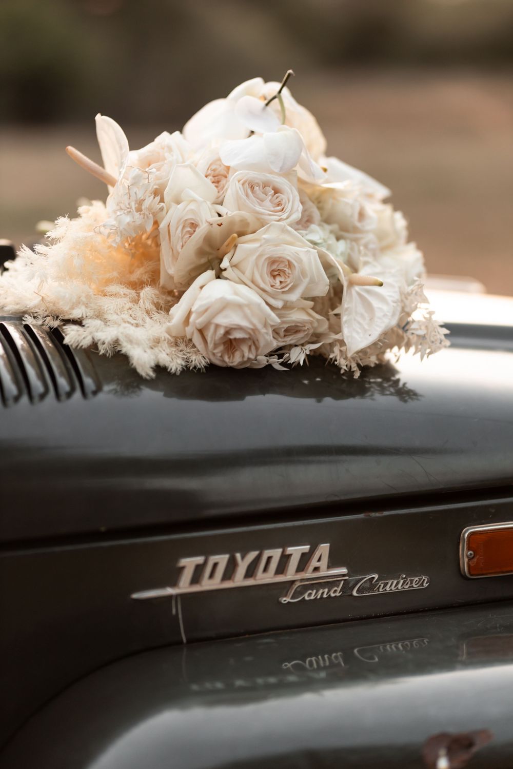 Bridal bouquet of cream roses resting on a vintage Toyota Land Cruiser hood at a wedding.