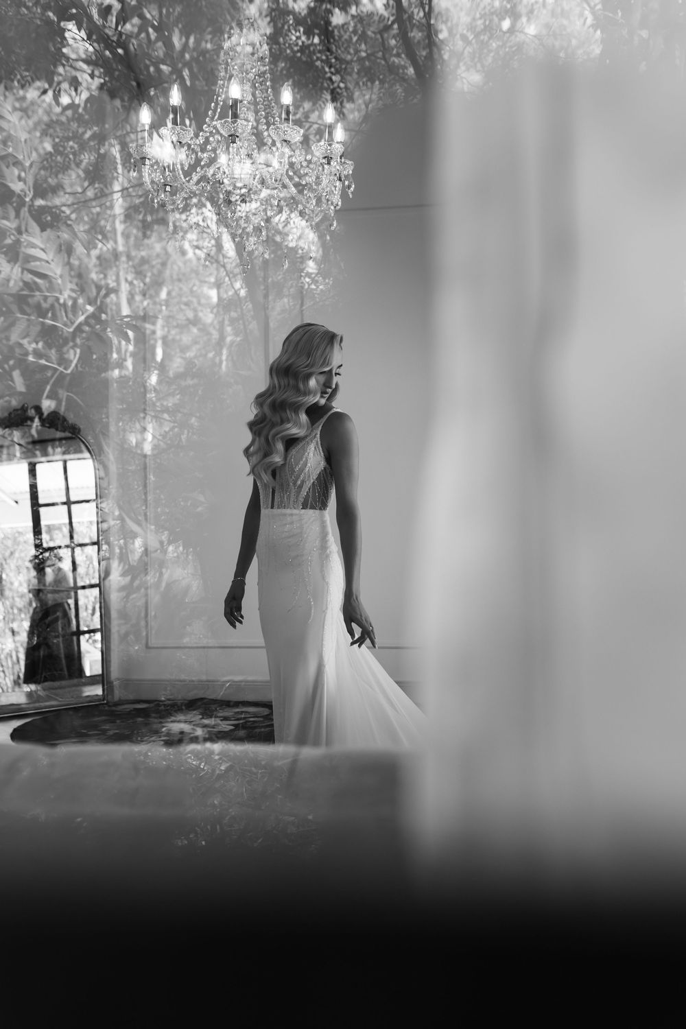 Elegant blonde bride in fitted wedding gown stands beneath crystal chandelier in dreamy black and white.