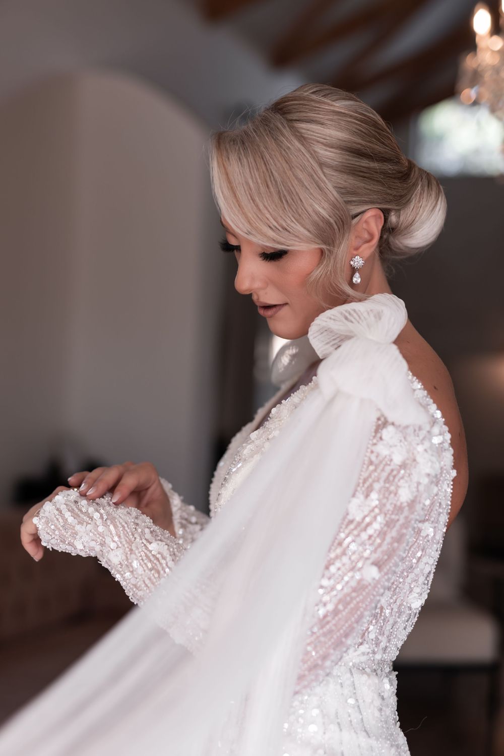 Elegant bride in sequined wedding gown with bow detail and updo hairstyle gazes down in soft indoor light.