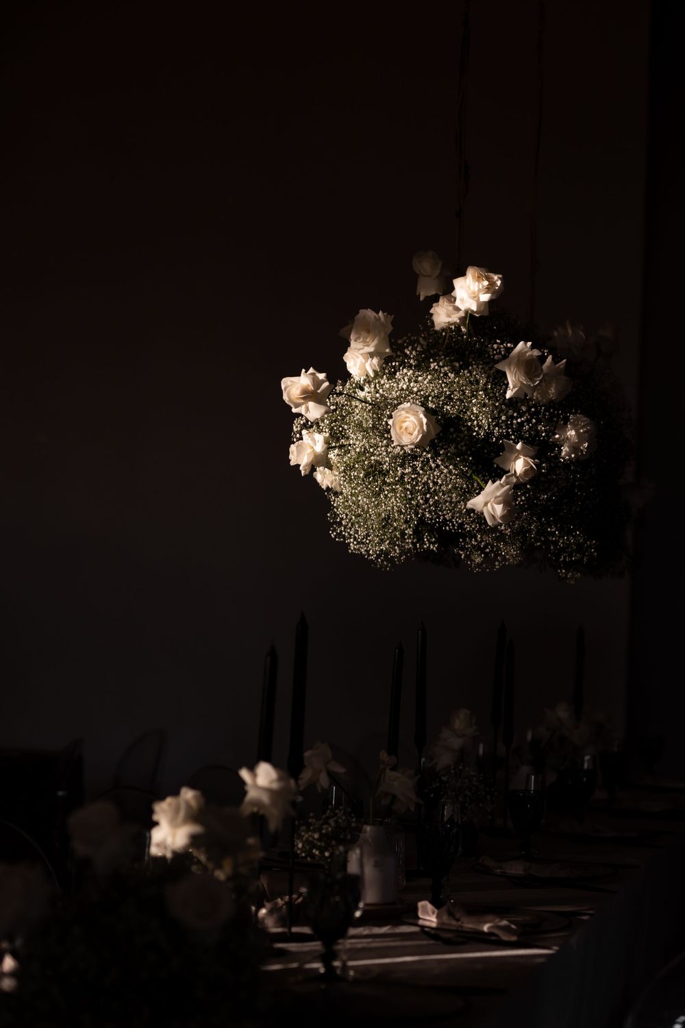 Elegant dark wedding table setting with glowing white roses and baby's breath floral chandelier above black candles.