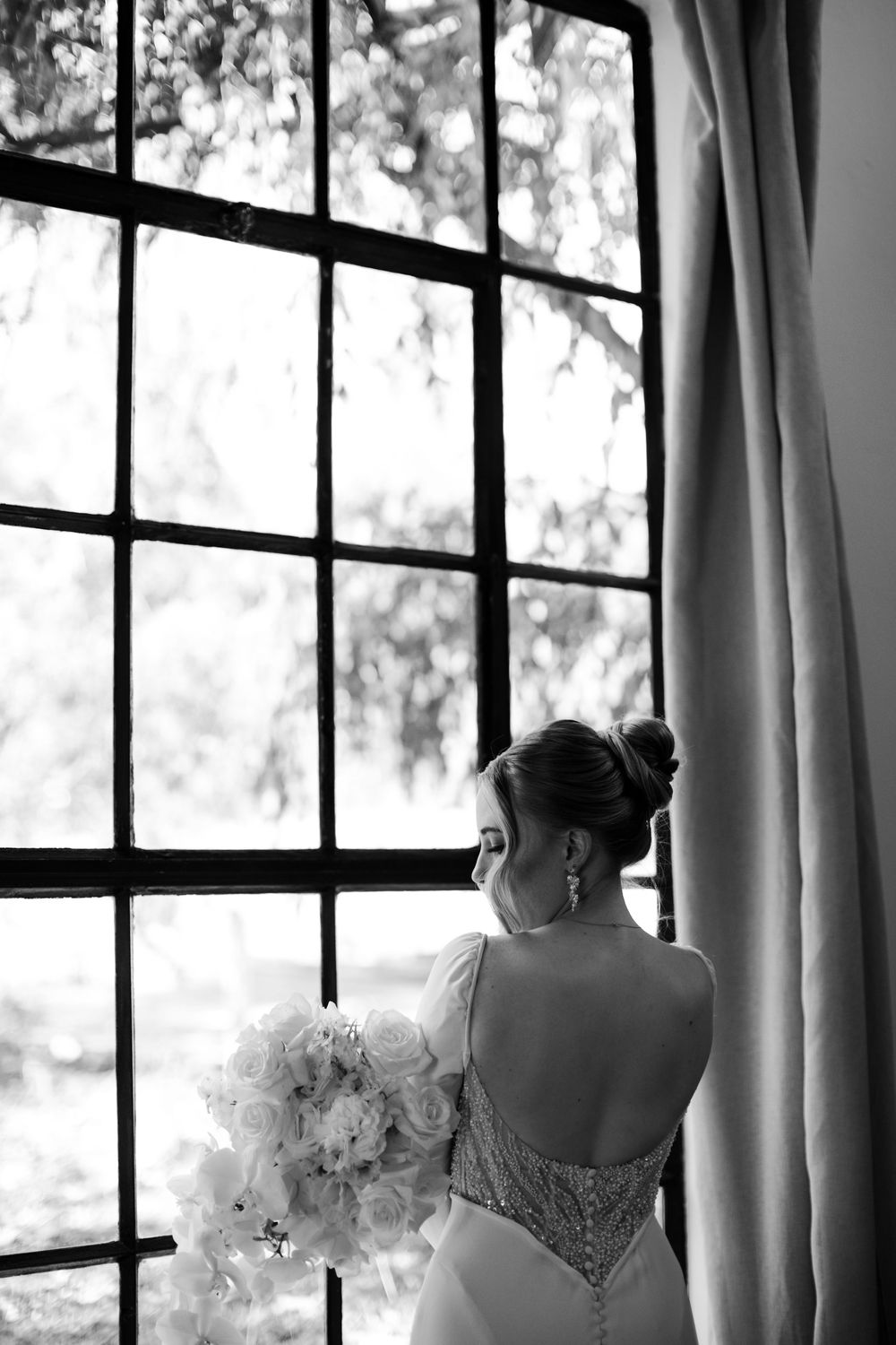 Bride in backless wedding gown holds white bouquet, gazing out large window in black and white photo.