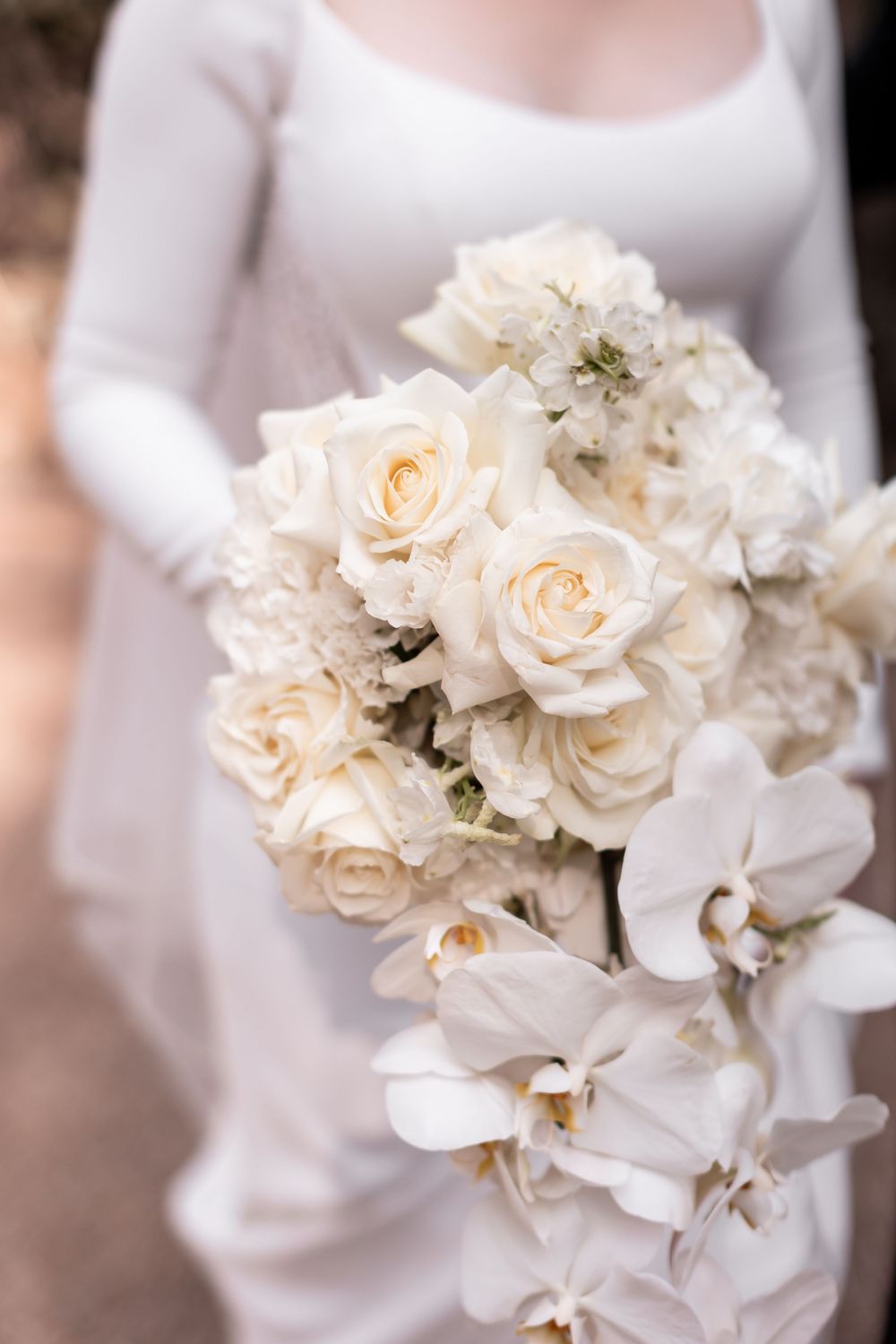 Bride holding lush white wedding bouquet of roses and orchids in cascading arrangement against white dress.