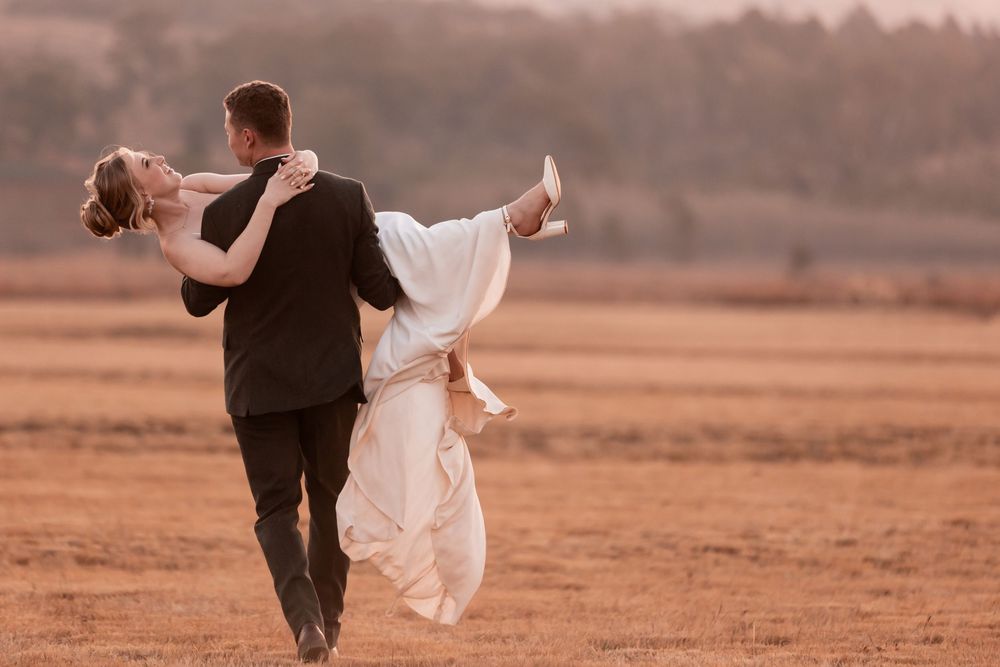 Groom lifting bride in white dress during romantic outdoor wedding photo shoot in an open field at sunset.