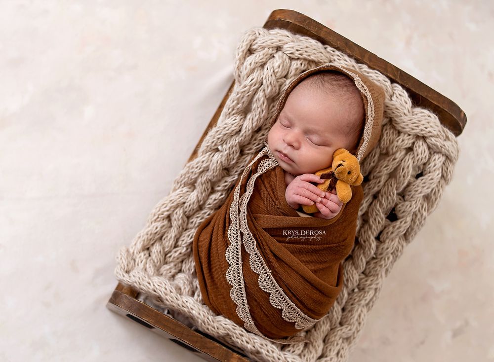 Newborn baby swaddled in rust wrap with tiny bear pacifier, sleeping peacefully in rustic wooden crate with cream knit blanket.