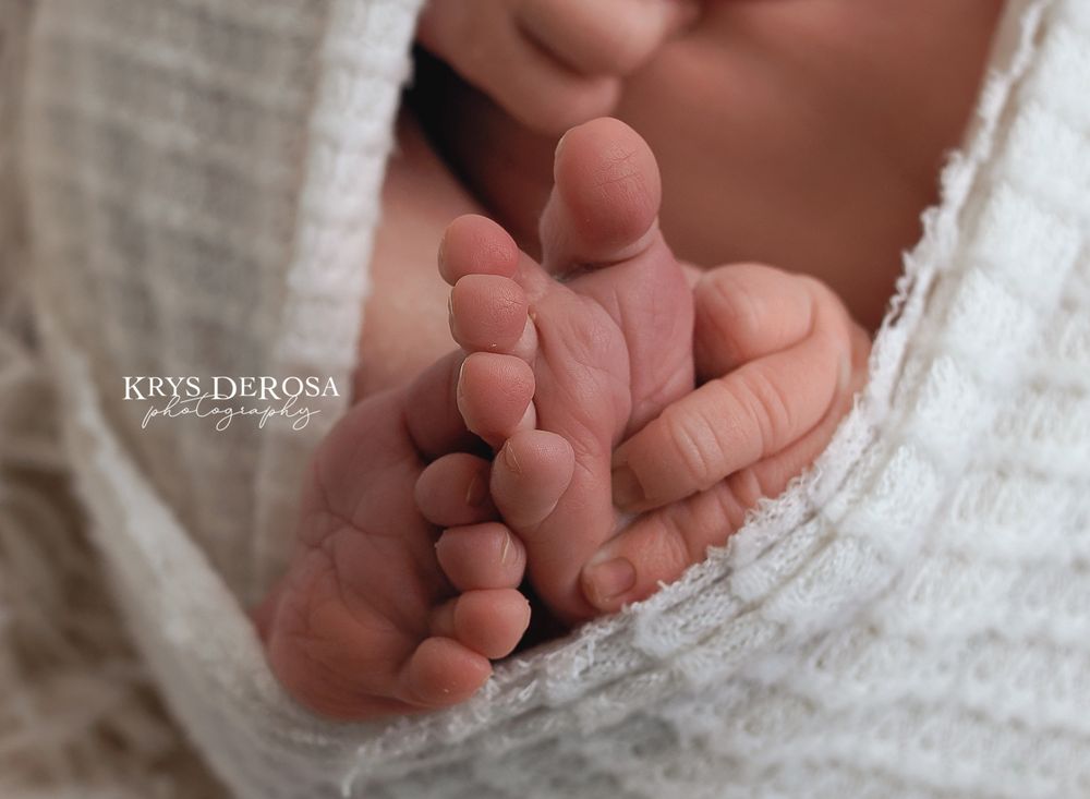 Close-up of tiny newborn baby feet wrapped in a soft white knit blanket, photographed by Krys DeRosa Photography.