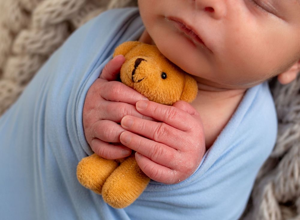Newborn baby wrapped in blue swaddle holding a small knitted yellow teddy bear, close-up photography.