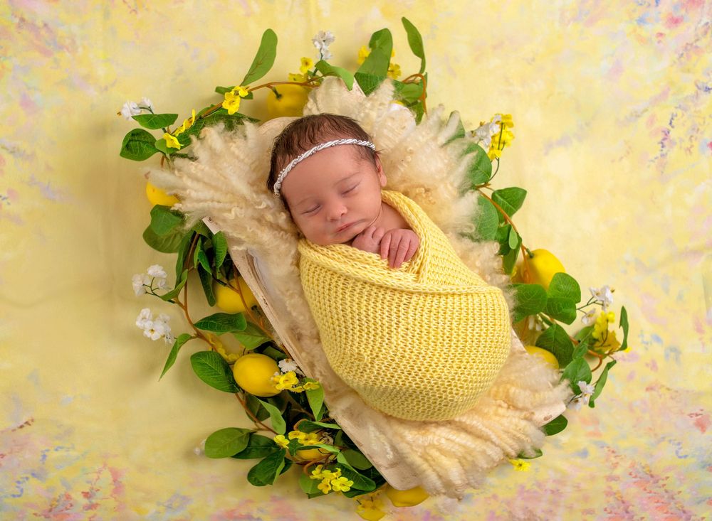 Newborn baby swaddled in yellow knit wrap sleeping in a floral basket with lemons and green leaves.