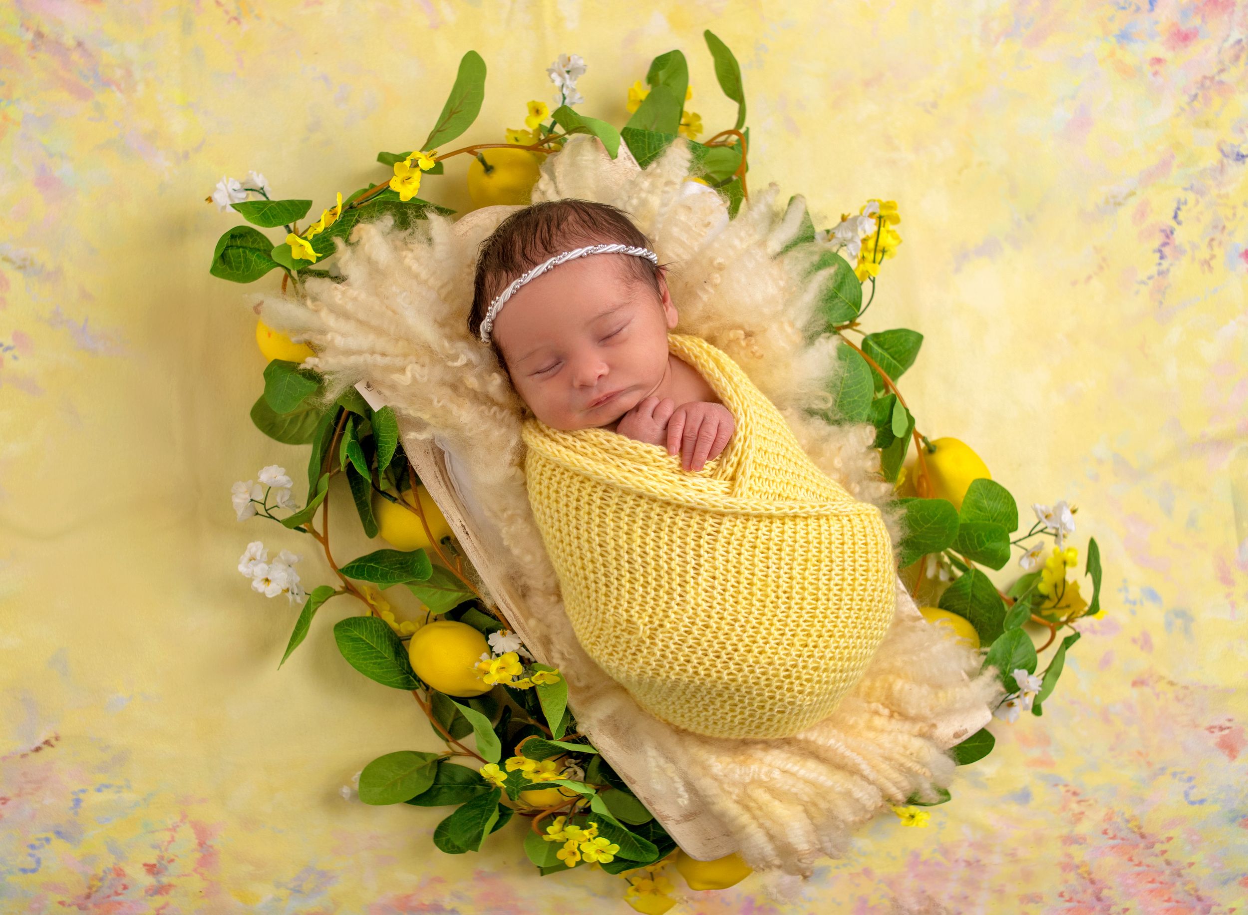 Newborn baby swaddled in yellow knit wrap sleeping in a floral basket with lemons and green leaves.