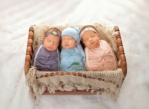 Three sleeping newborn triplets swaddled in purple, blue, and pink wraps with hats, nestled in a wicker basket.
