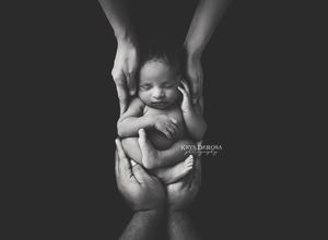 Black and white newborn photography of a sleeping baby cradled in parents hands against dark background.