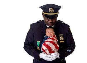 Police officer in dress uniform holding a sleeping baby wrapped in an American flag.
